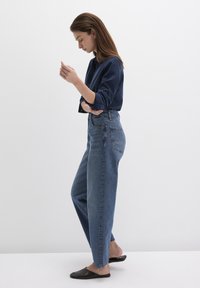High-waisted blue jeans in denim with a straight leg fit. Paired with a dark blue long-sleeve shirt and black slip-on shoes.