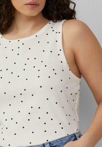 Mouwloze, ribgebreide top in crème met zwarte stippen. Heeft een ronde halslijn en een nauwsluitende pasvorm, gecombineerd met denimshorts.