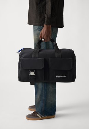 Black duffel bag with two front pockets, metallic clasp, logo patch, and blue zipper pull, made from smooth, durable fabric.