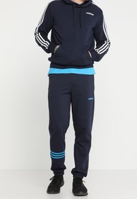 Námorně modrá mikina Adidas s bílými pruhy a logem, spárovaná s odpovídajícími tepláky s modrými akcenty a pruhy na kotnících. Černé tenisky.