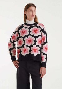 Mujer vestida con un suéter negro con grandes patrones de flores centrados en un corazón rosa y rojo, llevado sobre un cuello de tortuga blanco, combinado con pantalones negros.