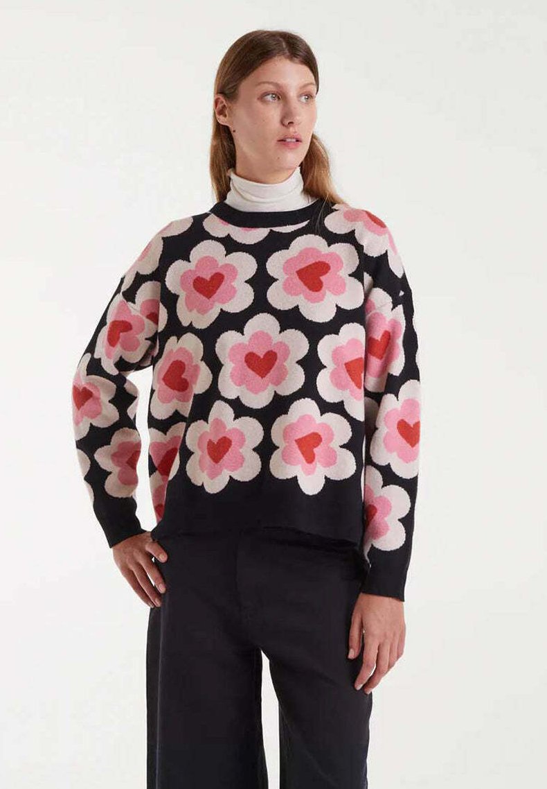 Mujer vestida con un suéter negro con grandes patrones de flores centrados en un corazón rosa y rojo, llevado sobre un cuello de tortuga blanco, combinado con pantalones negros.