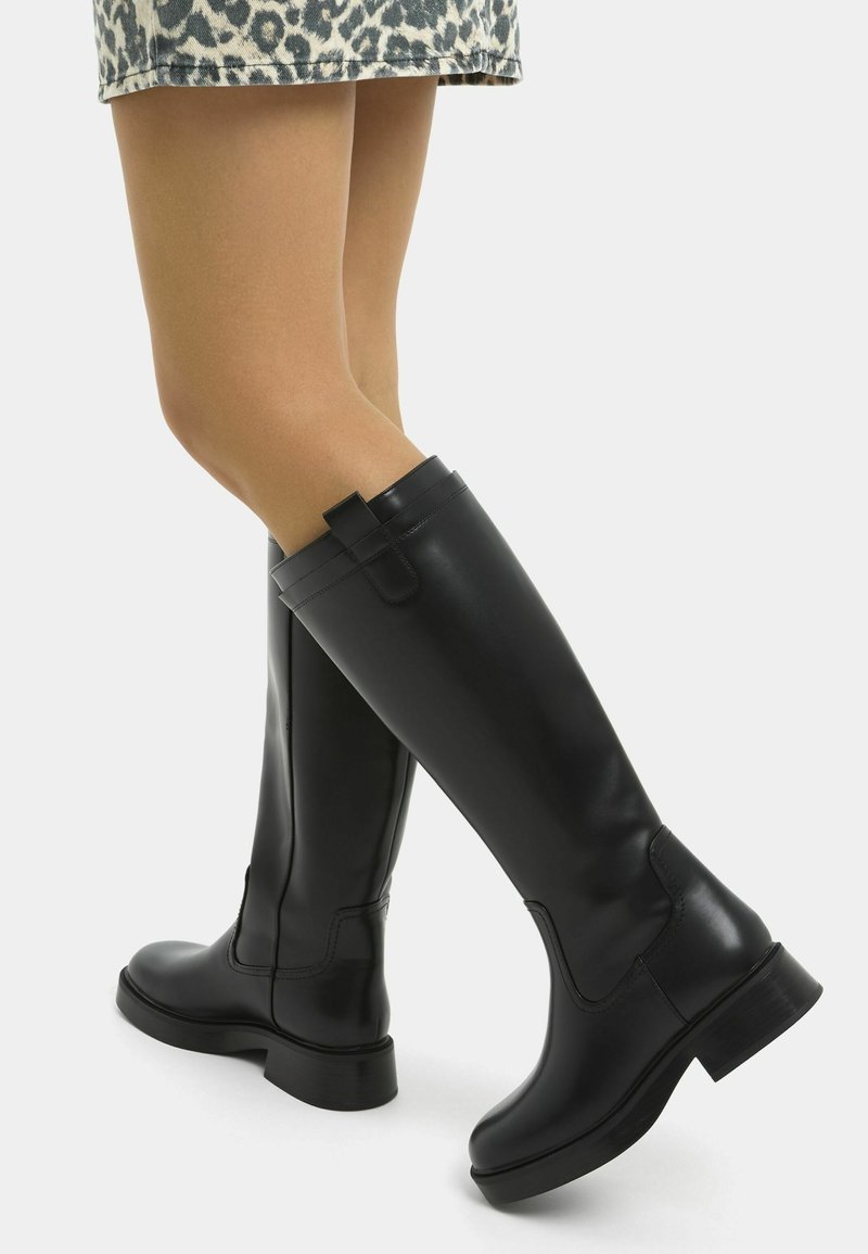 Piernas vistiendo botas de cuero negro hasta la rodilla y una falda corta de estampado de leopardo, con un pie ligeramente levantado, contra un fondo liso.