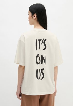 Kobieta z długimi czarnymi włosami nosi białą, oversize koszulkę z czarnym nadrukiem „IT'S ON US” pionowo na plecach.