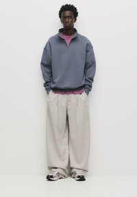 Felpa grigia in pile con zip a metà, sovrapposta a una maglietta rosa, abbinata a pantaloni beige larghi e sneakers multicolori.