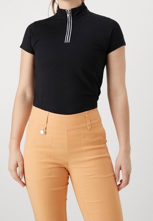 Vrouw draagt een zwart kort mouwen top met ritssluiting en beige hoge taille broek, staand tegen een eenvoudige lichte achtergrond.