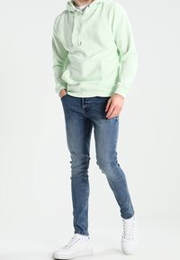 Persona che indossa un hoodie verde chiaro, jeans slim-fit blu e sneakers bianche, in piedi con una mano in tasca.
