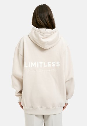 Person mit langen braunen Haaren, der einen übergroßen beigen Hoodie mit heruntergezogener Kapuze trägt, auf dessen Rückseite der Text "LIMITLESS" gedruckt ist. Sie steht vor einem weißen Hintergrund.