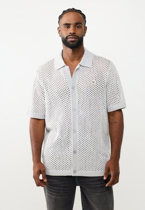 Man met gevlochten haar en baard staat recht vooruit, draagt een wit opengewerkt T-shirt met korte mouwen en een donkere jeans tegen een effen achtergrond.