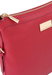 Rode leren clutch met een gouden rits en een gouden logo plaatje. Heeft een gladde textuur en een strakke, rechthoekige vorm.