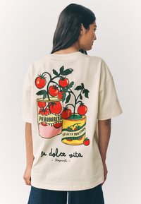 Bézs extra méretű póló színes illusztrációval, amely paradicsomnövényeket ábrázol konzervdobozokban, "Pomodorino" és "Italian Tomatoes" felirattal, valamint "la dolce vita Napoli" szöveggel.