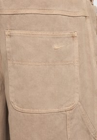 Beiga denimbyxor med en bakficka som har en broderad Nike-logotyp. Tyget har en mjuk textur med synliga sömdetaljer.