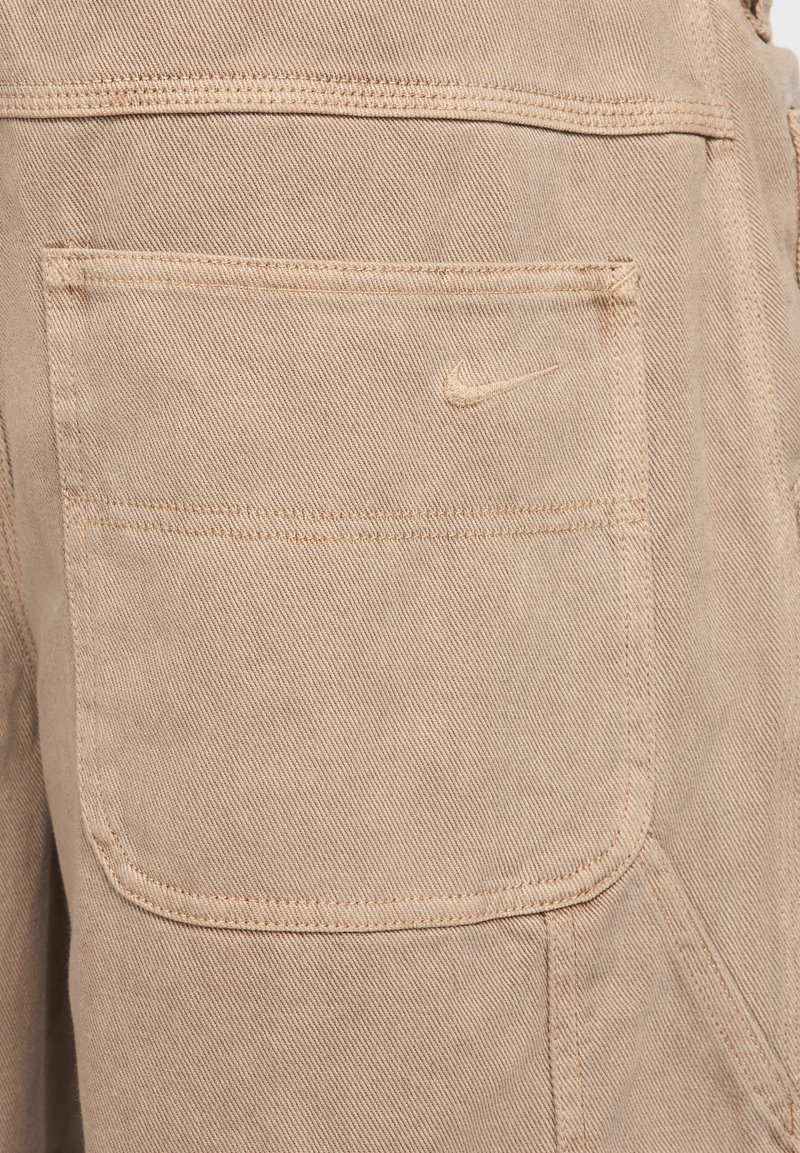 Beżowe dżinsy z tylną kieszenią z haftowanym logo Nike. Tkanina ma gładką teksturę z widocznymi detalami szwów.