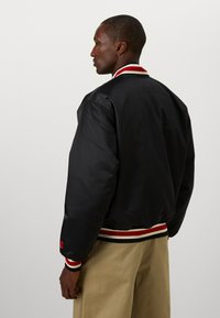 Black satin bomber jacket with striped red and white collar and cuffs, featuring a textured logo on the back and relaxed fit.