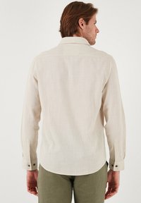 Camisa de manga larga beige claro con una tela texturizada y un cuello a medida. Presenta puños abotonados y un dobladillo trasero recto.