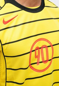 Žltý dres Nike s čiernymi horizontálnymi pruhmi. V strede sa nachádza okrúhly červený grafický prvok "90" a malá červená značka Nike na ľavom ramene.