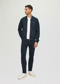 Námornícky zipsový sveter s rebrovanými manžetami, skombinovaný s tmavými nohavicami a bielymi šnurovacími topánkami. Minimalistický design, bez vzorov alebo akcentov.
