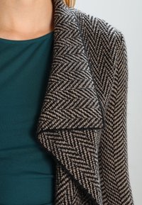 Strickjacke mit Schalkragen aus einer Wollmischung und einem Fischgrätmuster in Schwarz und Beige, die sanft über einem tealfarbenen Oberteil fällt. Strukturierter Stoff.