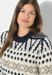 Neulepaita vaalean kermanvärisessä, laivastonsinisessä ja taupe-värisessä, jossa on geometrisia kuvioita; siinä on ribatty pyöreä pääntie ja teksturoitu kangas.