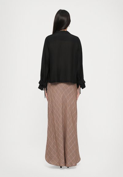 Woman with long dark hair wearing a sheer black blouse and a high-waisted, ankle-length brown plaid skirt, standing facing away.