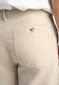 Pantalones beige de espiga con un bolsillo trasero de botón y una pequeña etiqueta. El material parece texturizado y transpirable.