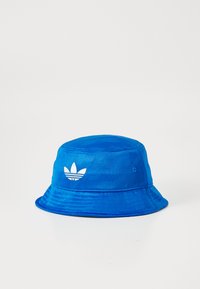 Mėlynas kibirėlis, pagamintas iš tekstūruoto audinio, su baltu Adidas logotipu priekyje ir plačiu,pliušiniu kraštu.