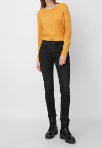 Pull jaune à manches longues, col rond, associé à un jean slim noir avec une fermeture à deux boutons. Modèle portant des bottes noires à lacets.