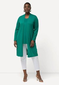 Turkusowy długi kardigan wykonany z miękkiego materiału, nałożony na pasującą bluzkę, w połączeniu z białymi spodniami do kostek i sandałami na obcasie.