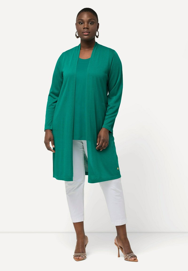 Turkusowy długi kardigan wykonany z miękkiego materiału, nałożony na pasującą bluzkę, w połączeniu z białymi spodniami do kostek i sandałami na obcasie.