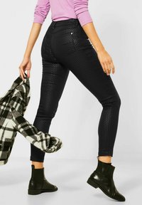 Černé skinny džíny s pruhem s texturovaným povrchem, středně vysokým pasem a dvěma zadními kapsami. Modelka drží kostkovanou bundu a má na sobě kotníkové boty.