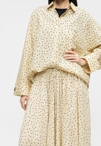 Chemise beige à boutons et jupe assortie avec des pois noirs, taille élastique avec cordon, portée sur un haut noir.