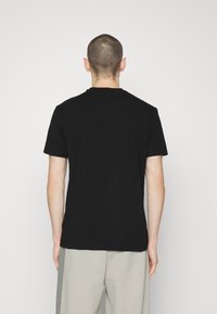 Camiseta de algodón negra, de manga corta, cuello redondo, diseño liso. Vista desde atrás, combinada con pantalones gris claro. Silueta simple y elegante.