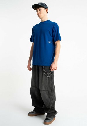 Młody mężczyzna w niebieskiej koszulce, luźnych czarnych spodniach z łańcuszkiem, szarych tenisówkach i czarnej czapce na jednolitym tle.