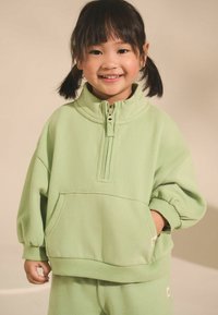 Vaaleanvihreä fleece-tyyppinen vetoketjullinen huppari, jossa on korkea kaulus, joustavat hihansuut ja kengurutasku. Yhdistetty yhteensopiviin housuihin.