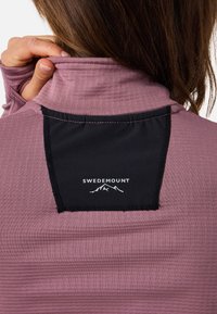 Persona che sistema una camicia a maniche lunghe color malva con toppa nera sulla parte superiore della schiena con la scritta "SWEDEMOUNT" e un logo montano.