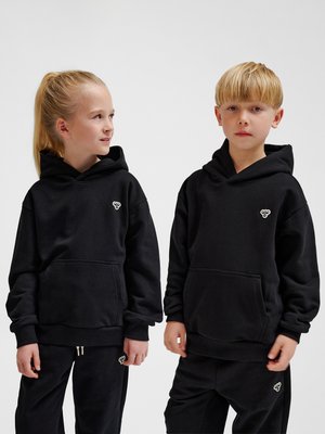 To børn iført matchende sorte hoodies og bukser med et lille hvidt logo, stående mod en ensfarvet lys baggrund.
