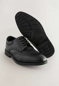 Chaussures brogue en cuir noir avec perforations décoratives, lacets et semelle en caoutchouc texturé, affichant un design classique.