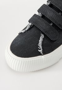 Mustat denimpüksid Velcro kinnitusrihmadega, kulunud tekstuur, valge kummist tallaga ja ümmarguse varbaga. Omab frängitud servi.