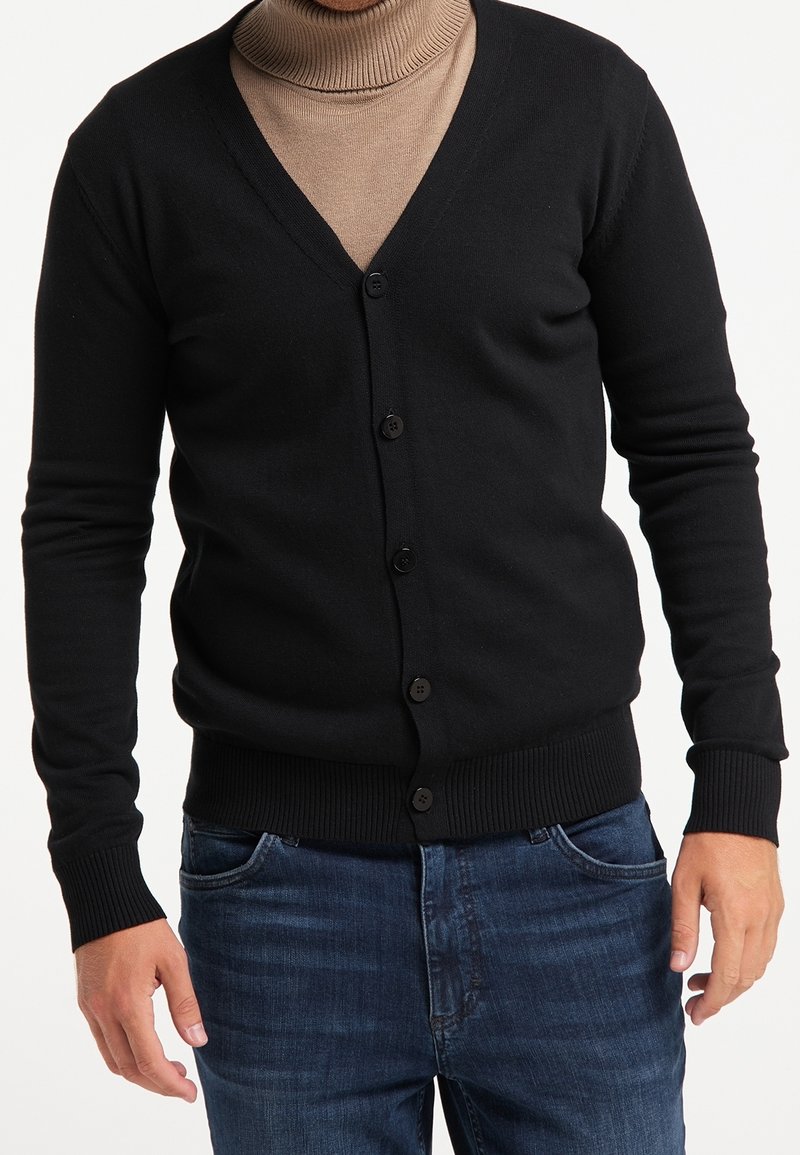 Uomo che indossa un cardigan nero con bottoni sopra un maglione col collo alto beige e jeans blu, in piedi con le mani rilassate ai fianchi.