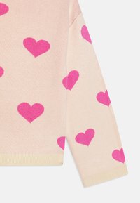 Pletený svetr z měkké růžové látky s velkými vzory srdcí v živé růžové. Žebrované manžety a lem s hladkou texturou. Pozorováno z bočního úhlu.