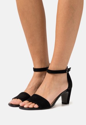 Des sandales à talons en suède noir présentent une large lanière au niveau des orteils, une lanière de cheville avec boucle et un talon bloc, offrant un design à la fois élégant et moderne.