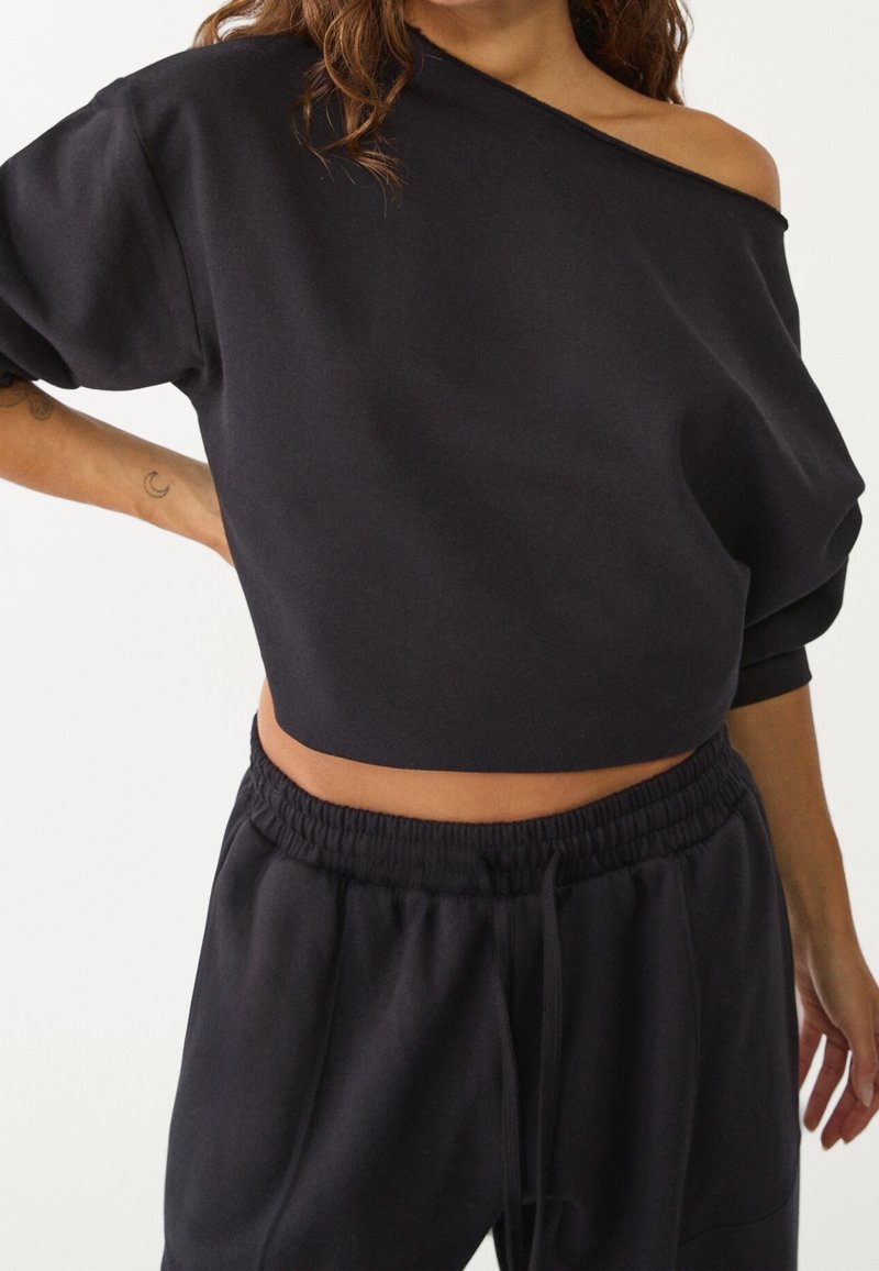 Femme portant un sweat court noir à épaules dénudées et un pantalon de survêtement noir assorti avec taille élastique.