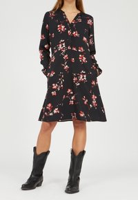 Vestido negro floral con mangas largas, con escote en V, bolsillos y un dobladillo acampanado, adornado con patrones florales en rosa y blanco. Llevado con botas negras.