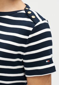 Μπλούζα σε ναυτικό μπλε και λευκές ρίγες, κατασκευασμένη από βαμβάκι· διαθέτει χρυσά κουμπιά στον ώμο και μικρό λογότυπο με κόκκινο, λευκό και μπλε χρώμα στο μανίκι.