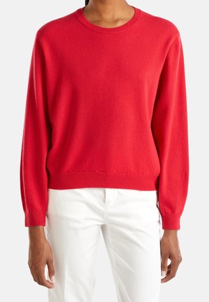 Personne portant un pull col rond rouge vif à manches longues associé à un pantalon blanc, debout sur un fond uni.