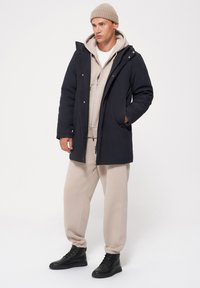 Granatowy parka z kapturem, z zapięciem na zamek i bocznymi kieszeniami, nałożona na beżowy hoodie i dresy, zestawiona z czarnymi butami.