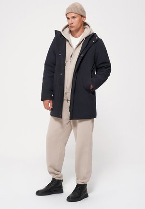 Granatowy parka z kapturem, z zapięciem na zamek i bocznymi kieszeniami, nałożona na beżowy hoodie i dresy, zestawiona z czarnymi butami.