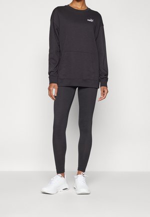 Person trägt schwarzen Puma-Sweatshirt mit Fronttasche, schwarze Leggings und weiße Sportschuhe, steht vor einem einfarbigen hellen Hintergrund.