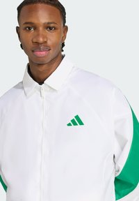 Giacca bianca con zip, dettagli verdi e logo Adidas verde. Presenta un colletto e una texture di tessuto liscia e elegante.