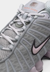 Κοντινό πλάνο γκρι αθλητικού παπουτσιού Nike Shox με λευκά κορδόνια, επάνω μέρος από δίχτυ, ροζ λογότυπο Nike και ορατές ελατηριωτές στήλες στη σόλα.